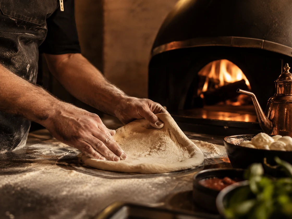 Chef stretching pizza dough by hand beside the glowing wood-fired oven